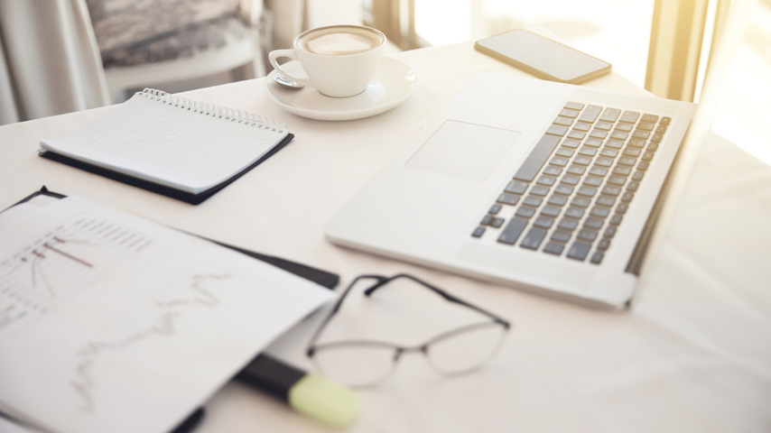 Sobre una mesa, un ordenador, libreta, gafas, taza de café,  rotuladores